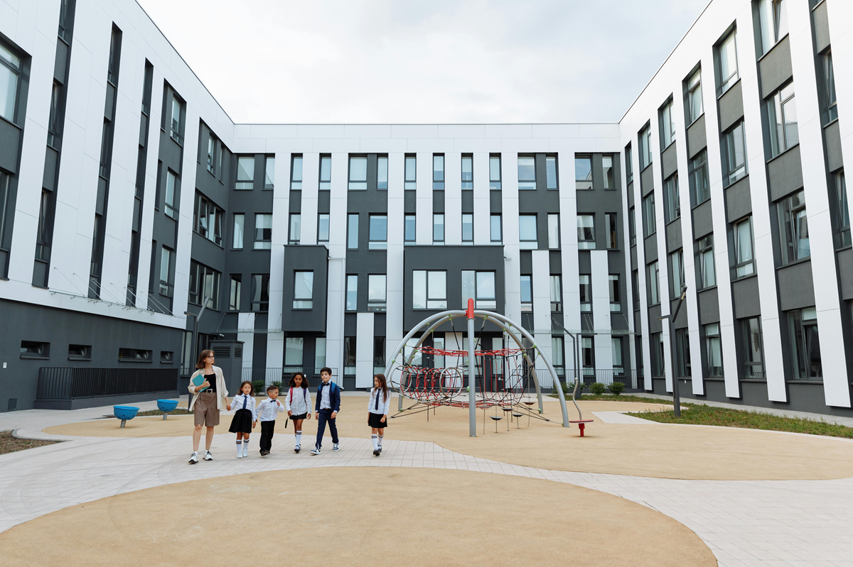 Modern school courtyard with a playground and students walking with a teacher in front of contemporary educational buildings.